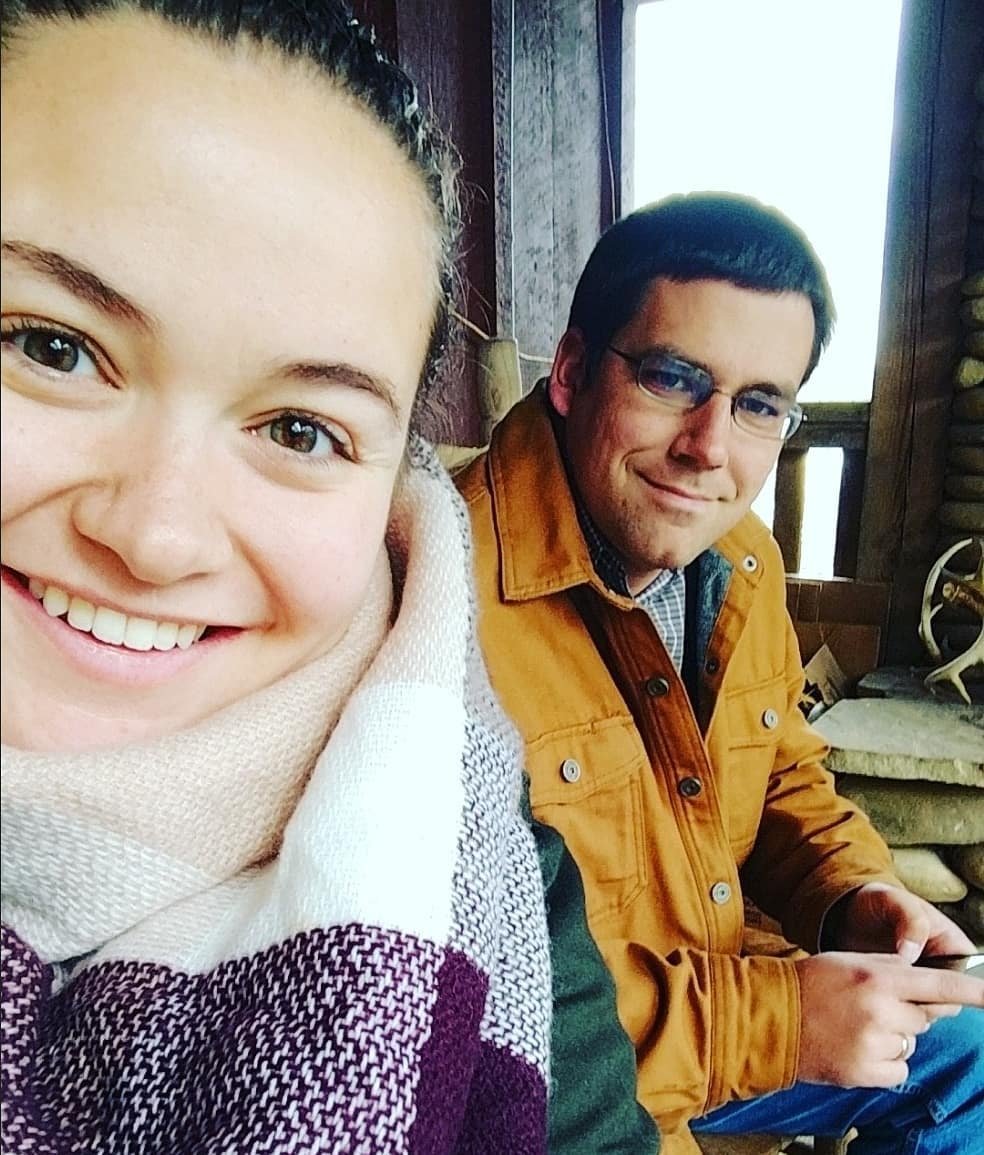 Young woman smiling in a cozy scarf poses for a selfie with a man sitting beside her, both enjoying a relaxed moment indoors. The background features wooden elements and natural light, creating a warm atmosphere.