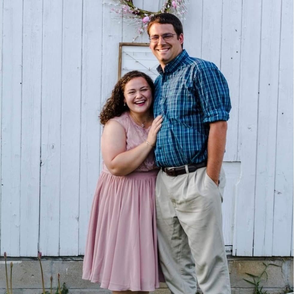 Happy couple posing together outdoors in front of a white wooden barn, with the woman wearing a pink dress and the man in a blue plaid shirt and beige pants, both smiling and enjoying their time together.