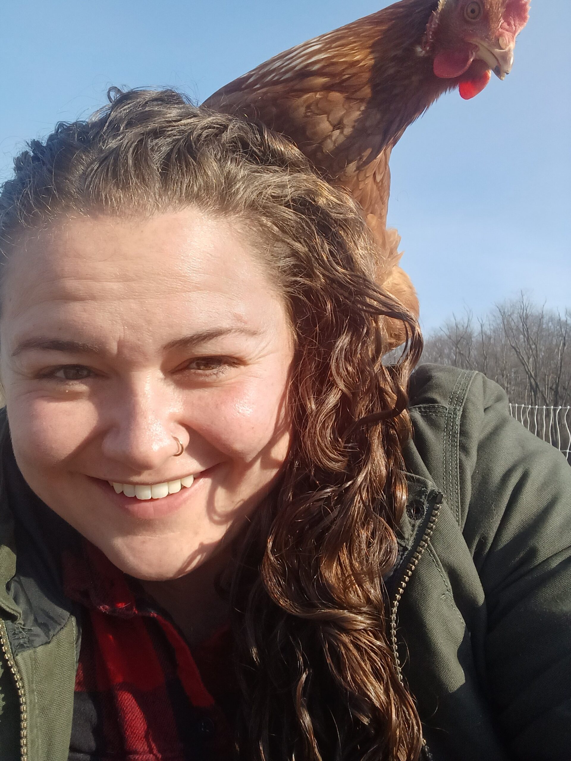 Woman smiling for a selfie with a chicken perched on her shoulder, set against a clear blue sky and a rural background.