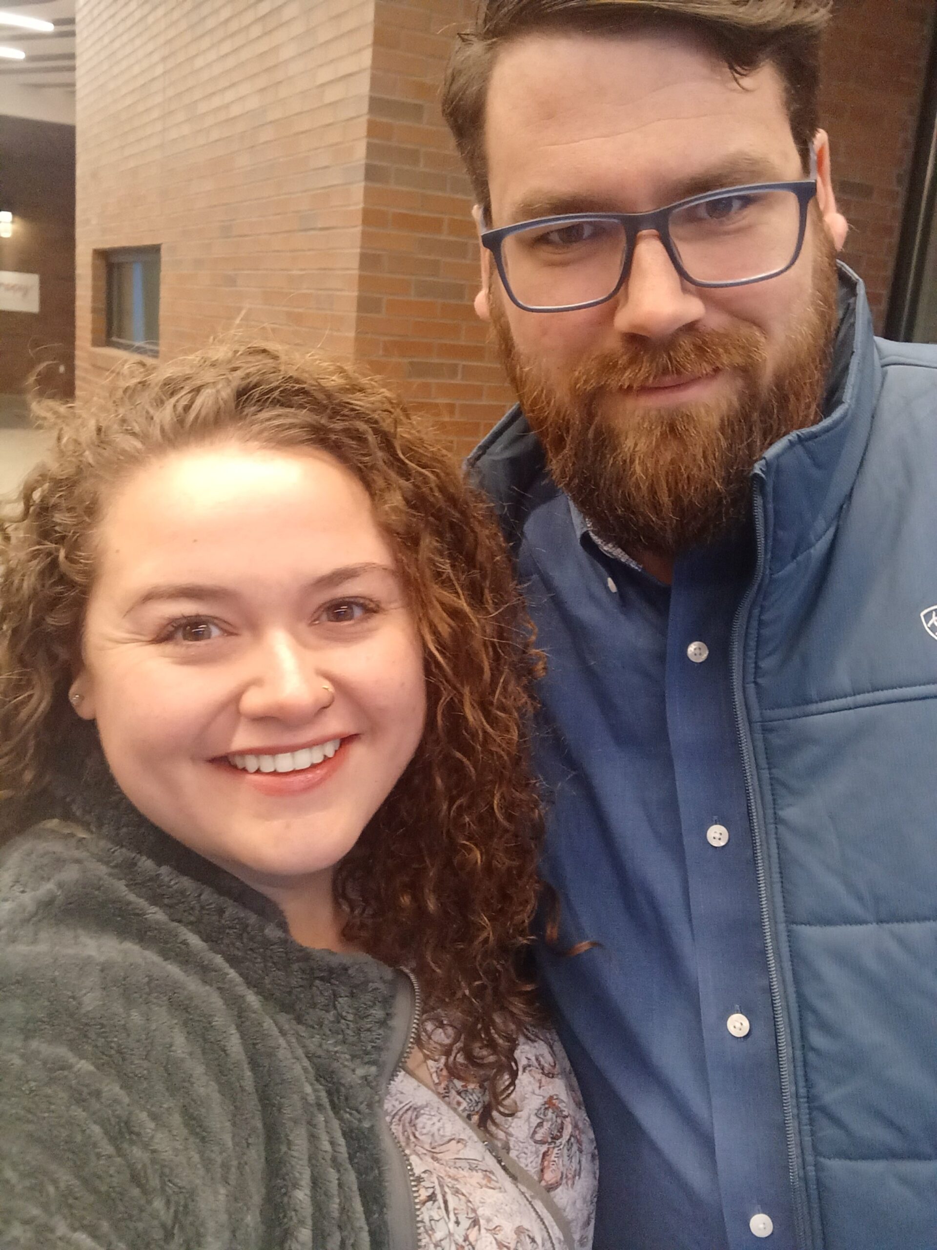 Two smiling individuals pose for a selfie in a brick-lined hallway. The woman has curly hair and is wearing a cozy grey jacket, while the man has a beard and glasses, dressed in a blue shirt and jacket. The background features a modern interior with soft lighting.