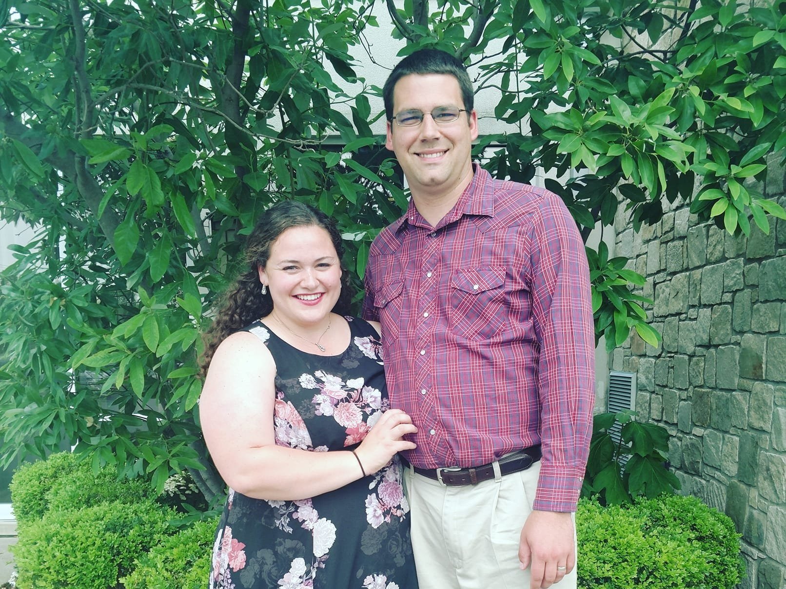 Smiling couple posing together outdoors in front of lush green foliage, the woman wearing a floral dress and the man in a plaid shirt, showcasing a joyful moment.