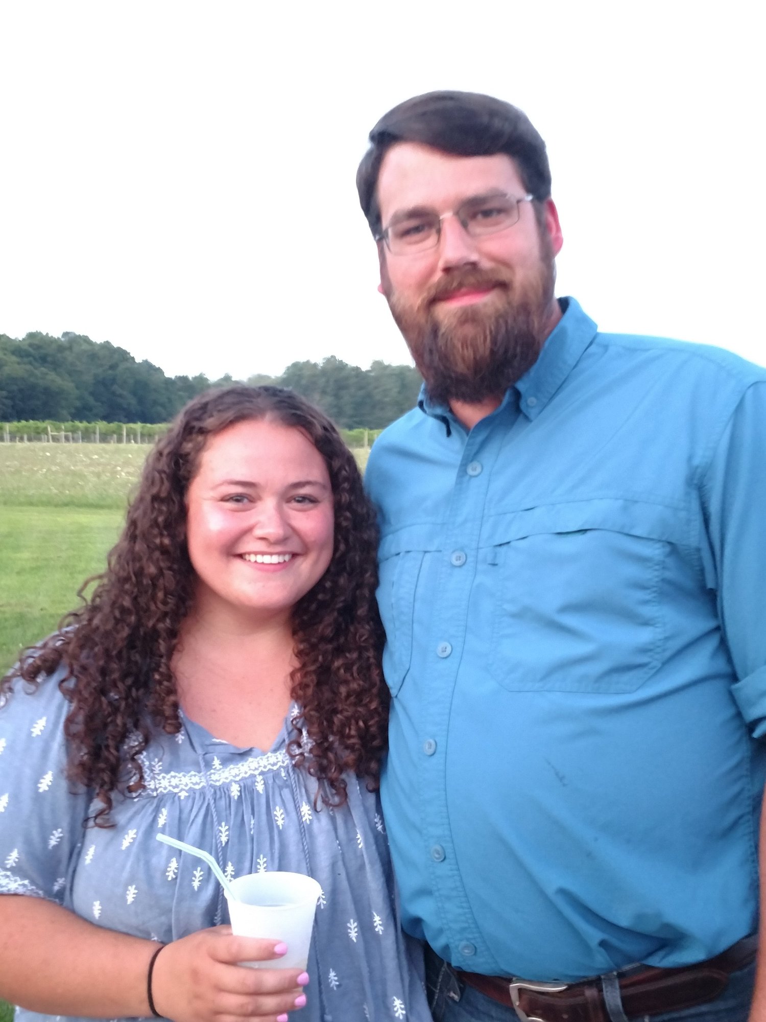 Happy couple posing outdoors, smiling together at a social event. The woman has curly hair and holds a drink, while the man wears glasses and a blue shirt. A green landscape with trees is visible in the background.