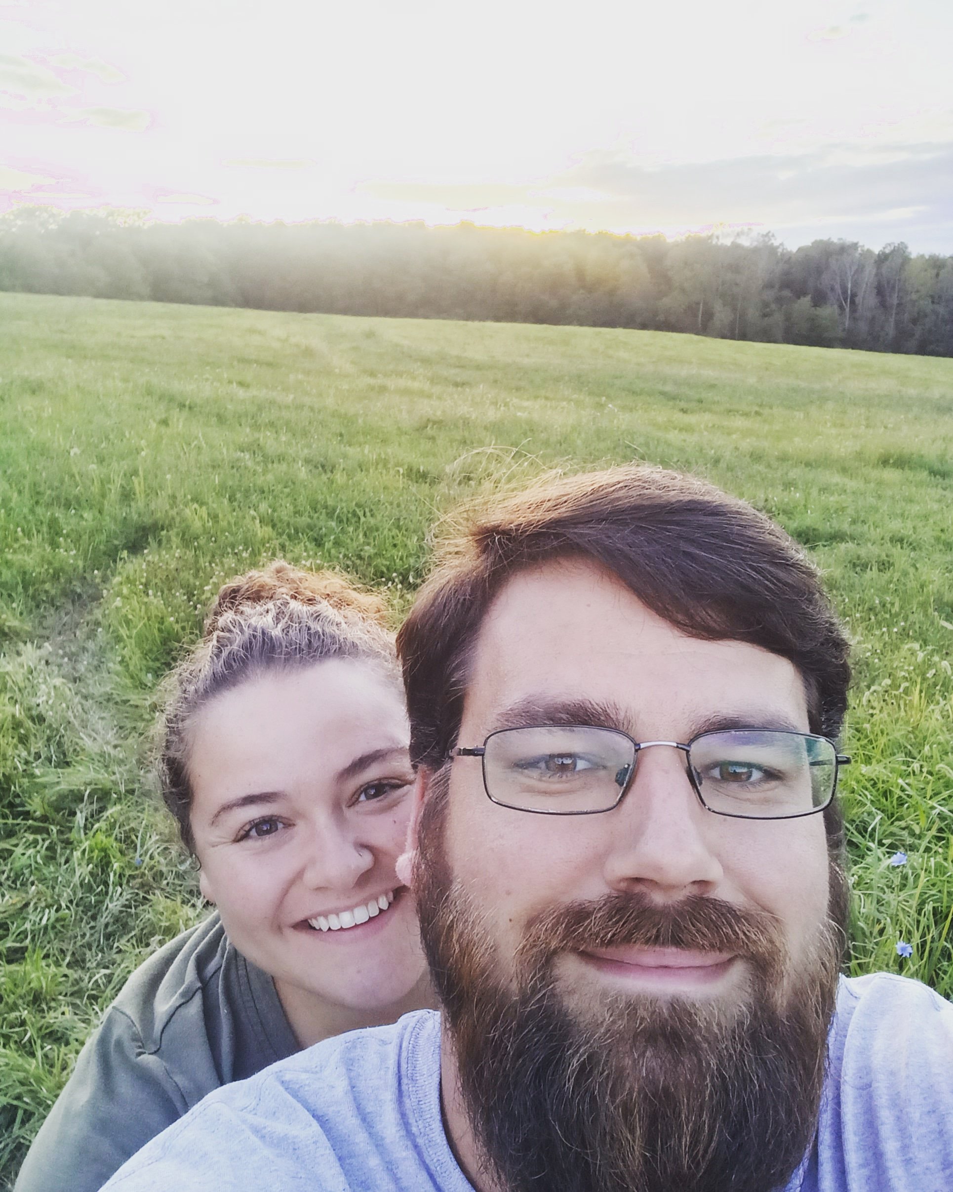 Couple smiling together in a lush green field at sunset, showcasing a romantic outdoor moment surrounded by nature.