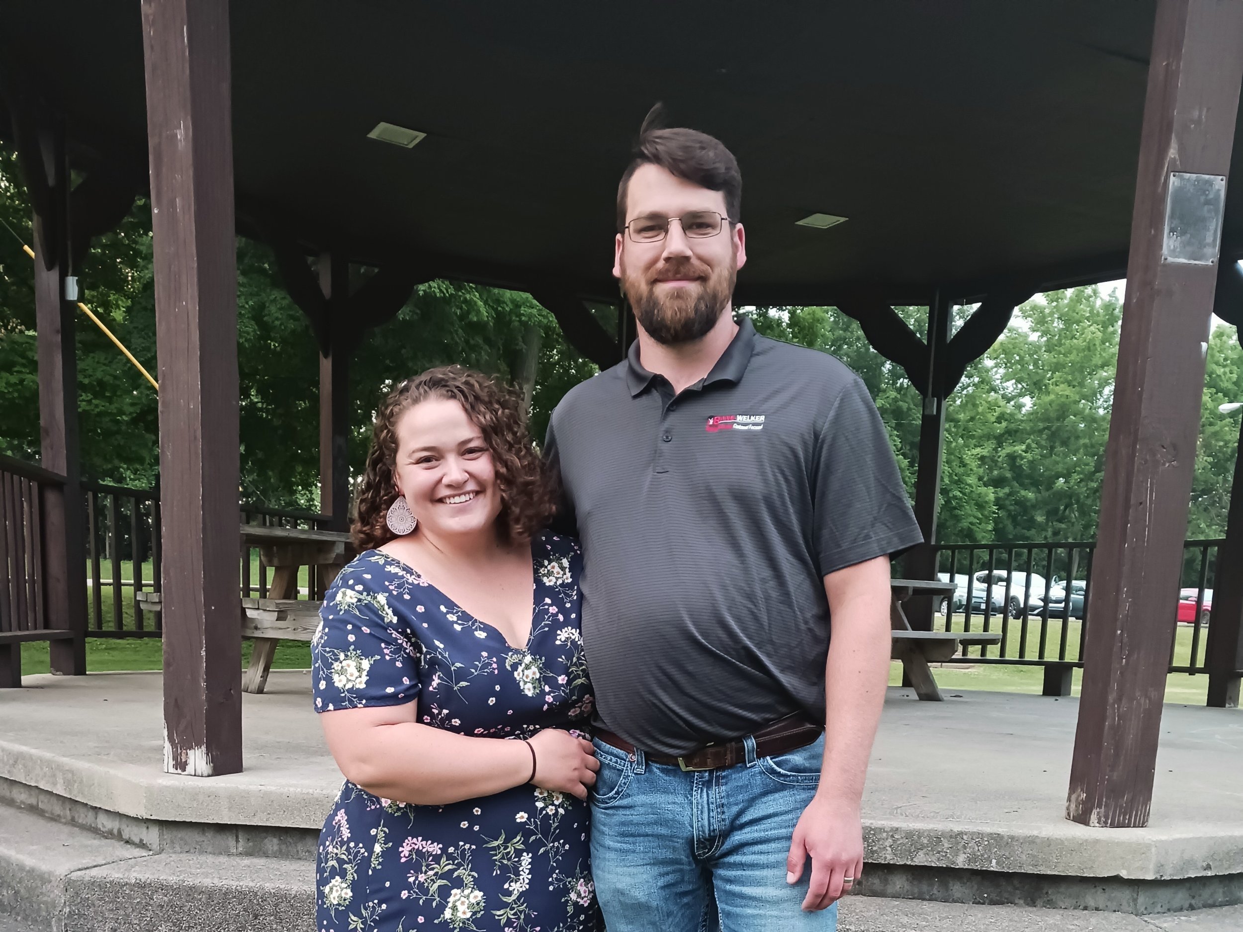 Smiling couple standing together under a gazebo in a park, with green trees in the background. The woman is wearing a floral dress and the man is dressed in a polo shirt and jeans.