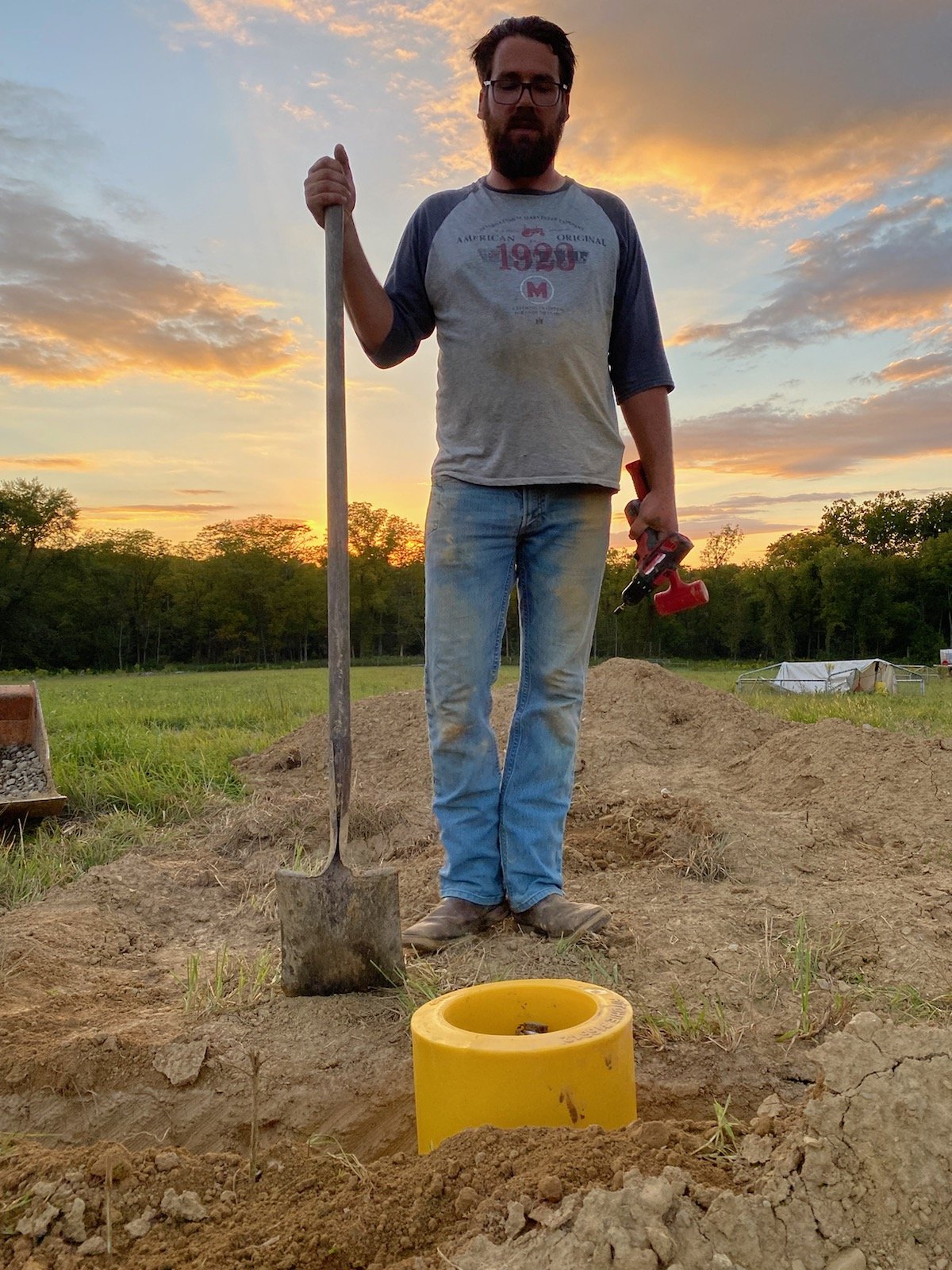 Man holding a shovel and a cordless drill, standing next to a yellow pipe fitting in a dug hole, with a sunset and green field in the background. Ideal for topics on landscaping or DIY projects.